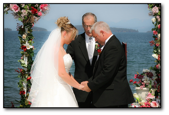 Bride and groom bowed in prayer in front of minister on the shore of a mountain lake.